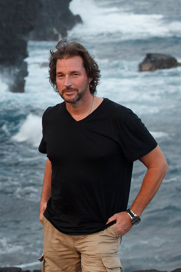 Man standing in front of ocean in black shirt and khaki shorts looking at camera